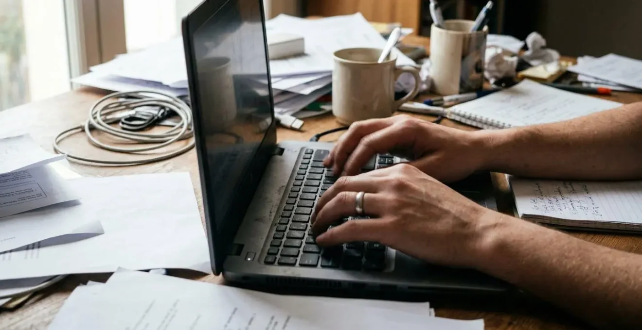 Close-up of hands typing on a laptop keyboard at a casual desk with papers and a smartphone visible