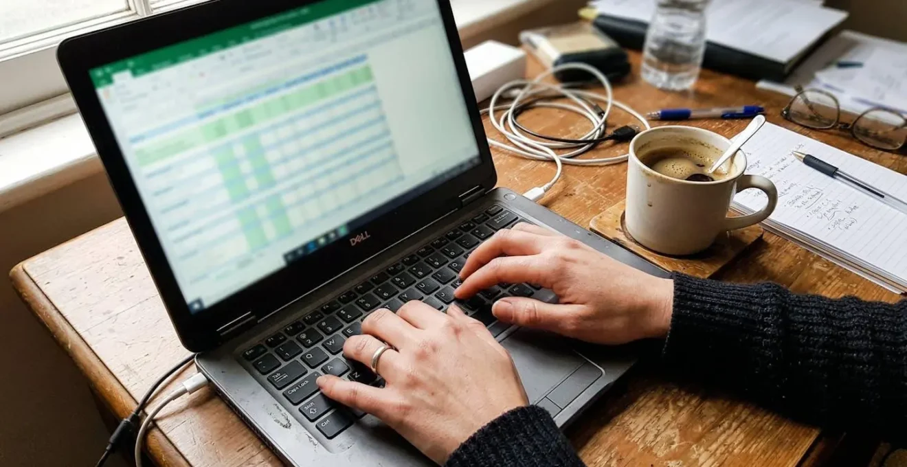 Hands resting near a laptop keyboard with a coffee cup beside it on a cluttered desk in natural daylight