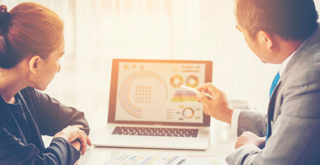 Team gathered around a conference table with one person gesturing toward a wall-mounted display in mixed office lighting